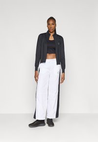 Černá zipová bunda přes černý crop top, doplněná bílými širokými kalhotami s černými bočními panely. Černé boty dokončují celý outfit.