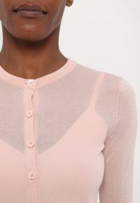 Femme portant un cardigan boutonné transparent rose pâle sur un caraco rose assorti, gros plan sur le haut du torse et l'encolure.