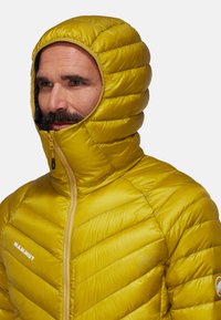 Veste isolante jaune avec une capuche ajustée, motif matelassé diagonal, fermeture éclair à l'avant et logo sur la poitrine. Texture légère et douce.