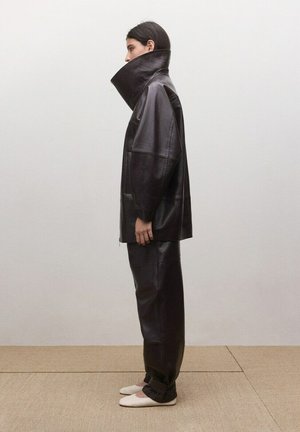 Persoană în profil lateral purtând o jachetă supradimensionată din piele maro închis cu guler înalt, pantaloni asortați din piele și pantofi albi cu talpă plată.