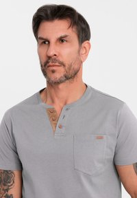 Šedé tričko s krátkým rukávem a Henley výstřihem, se třemi hnědými knoflíky, béžovým akcentem na krku a kapsou na hrudi. Měkká, hladká tkanina.