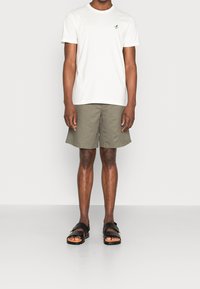 Hvid bomulds-t-shirt med lille grøn logo, parret med olivengrønne shorts og sorte sandaler med to stropper og en kraftig sål.