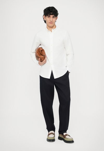 Biela košeľa na gombíky, tmavé nohavice, hnedá kožená taška a viacfarebné topánky. Košeľa má golier a je starostlivo zastrčená.