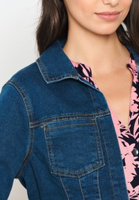 Jeansjacka i mörkblått med krage och bröstficka. Under den en rosa topp med blommönster och V-ringning.