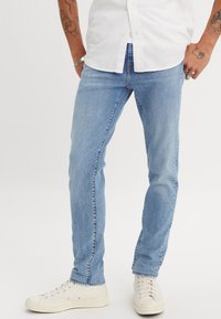 Világoskék farmernadrág, slim fit, egyenes lábvonallal és minimális kopásokkal, fehér vászon sportcipővel párosítva.