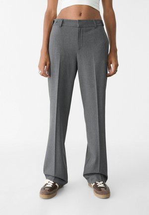 Volwassene draagt hooggetailleerde grijze wijde broek en bruine sneakers met witte veters, staand tegen een effen witte achtergrond.