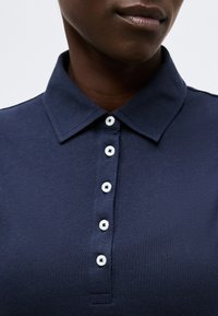 Κοντινό πλάνο ενός ατόμου με σκουρόχρωμο δέρμα που φοράει σκούρο μπλε πουκάμισο με γιακά και πέντε άσπρα κουμπιά κουμπωμένα μπροστά.