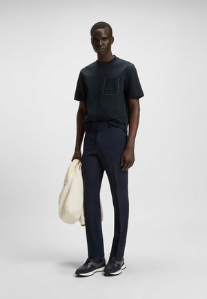Schwarzes Kurzarmshirt mit seitlichen Nähte und Brusttasche, kombiniert mit taillierten dunklen Hosen und strukturierten Sneakern. Leichte cremefarbene Jacke wird gehalten.