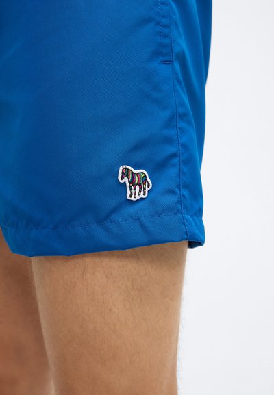 Siniset uimashortsit, jotka on valmistettu kevyestä materiaalista. Vasemmassa reunassa pieni kirjailtu monivärinen seepra-kuvio.