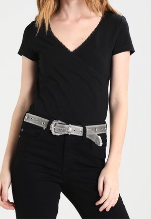 Femme portant un haut noir à manches courtes en V et un pantalon noir avec une ceinture argentée avec boucle décorative et œillets.