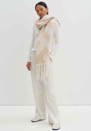 Beige wool scarf with geometric pattern and fringed edges, worn with a white sweater and light cream trousers. Smooth texture, medium thickness.