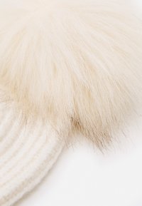 White knitted beanie with a large, fluffy faux fur pom-pom at the top. The texture is soft and the color is a light cream.
