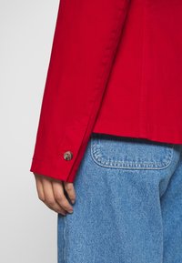Červená bavlněná bunda s knoflíkovým manžetou a viditelným švem, spárovaná se světle modrými džíny s zadní kapsou.