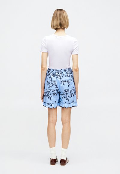 Γυναίκα με ξανθά κοντά μαλλιά, που φοράει λευκό κοντομάνικο πουκάμισο και ανοιχτό μπλε σορτς με λουλουδάτο σχέδιο και καμπυλωτές άκρες, λευκές κάλτσες και καφέ παπούτσια.