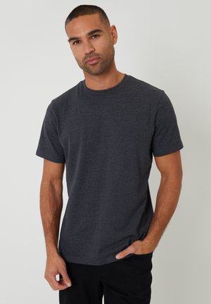 Mand med kort hår iført en mørkegrå t-shirt med korte ærmer og sorte bukser, stående med den ene hånd i lommen mod en ensfarvet baggrund.