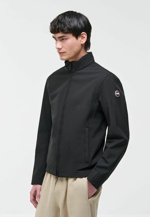 Młody mężczyzna ubrany w czarną kurtkę zapinaną na zamek z okrągłym logo na lewym rękawie i beżowe spodnie, stojący z rękami w kieszeniach.