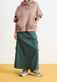 Ljusbrun oversized hoodie med en framficka, kombinerad med en mörkgrön lång cargojacka med sidofickor, samt grå löparskor.