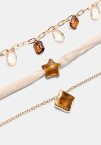Trois bracelets délicats avec des chaînes en or, des charms facettés marron et transparents, une perle en forme d'étoile marron sur tissu et un charm carré doré brillant.