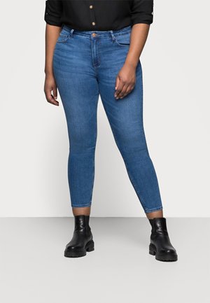 Person iført mellemblå skinny jeans, sort langærmet skjorte og sorte ankelstøvler, der står mod en hvid baggrund.