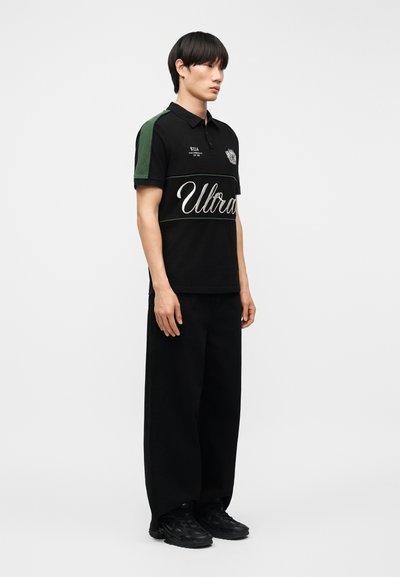 Νέος άνδρας που στέκεται, φοράει μαύρο πόλο με πράσινα μανίκια και άσπρο κείμενο "Ultra," συνδυασμένο με φαρδιά μαύρα παντελόνια και μαύρα παπούτσια.