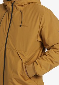 Chaqueta con capucha Quiksilver en color mostaza. Presenta un cierre de cremallera, cordón ajustable y puños elásticos; textura de tela suave.