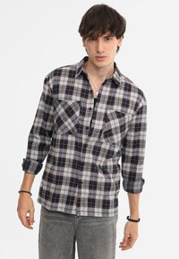 Camicia di flanella a quadri nei colori blu navy, grigio e rosso. Presenta due tasche sul petto, chiusura con bottoni e maniche arrotolate. Indossata con jeans grigi.