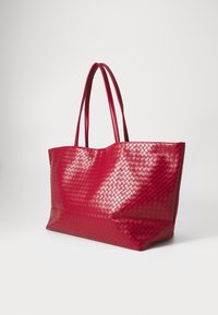 Sac fourre-tout tissé rouge avec une finition brillante, doté de deux longues poignées et d'une forme rectangulaire spacieuse. Motif texturé en losanges sur la surface.