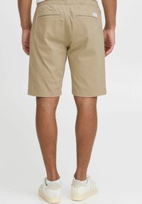 Beige shorts dengan pinggang elastis, dua saku belakang, dan potongan lurus, dipadukan dengan sepatu putih dan kaus kaki putih.