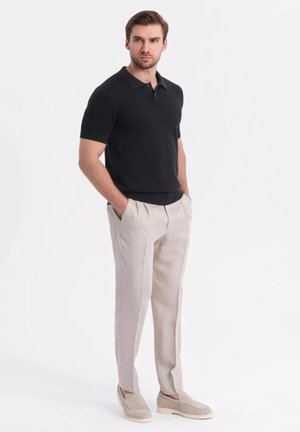 Sort kortærmet polo-shirt med krave, parret med lyse beige plisserede bukser og beige slip-ons, mod en hvid baggrund.