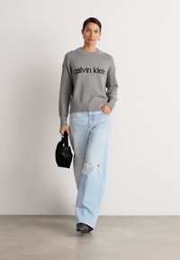 Hallitusseente mustriga hall kudum, millel on "calvin klein" tekst, sinised rebenenud laiad püksid ja väike must kott, modell seisab heleda tausta ees.