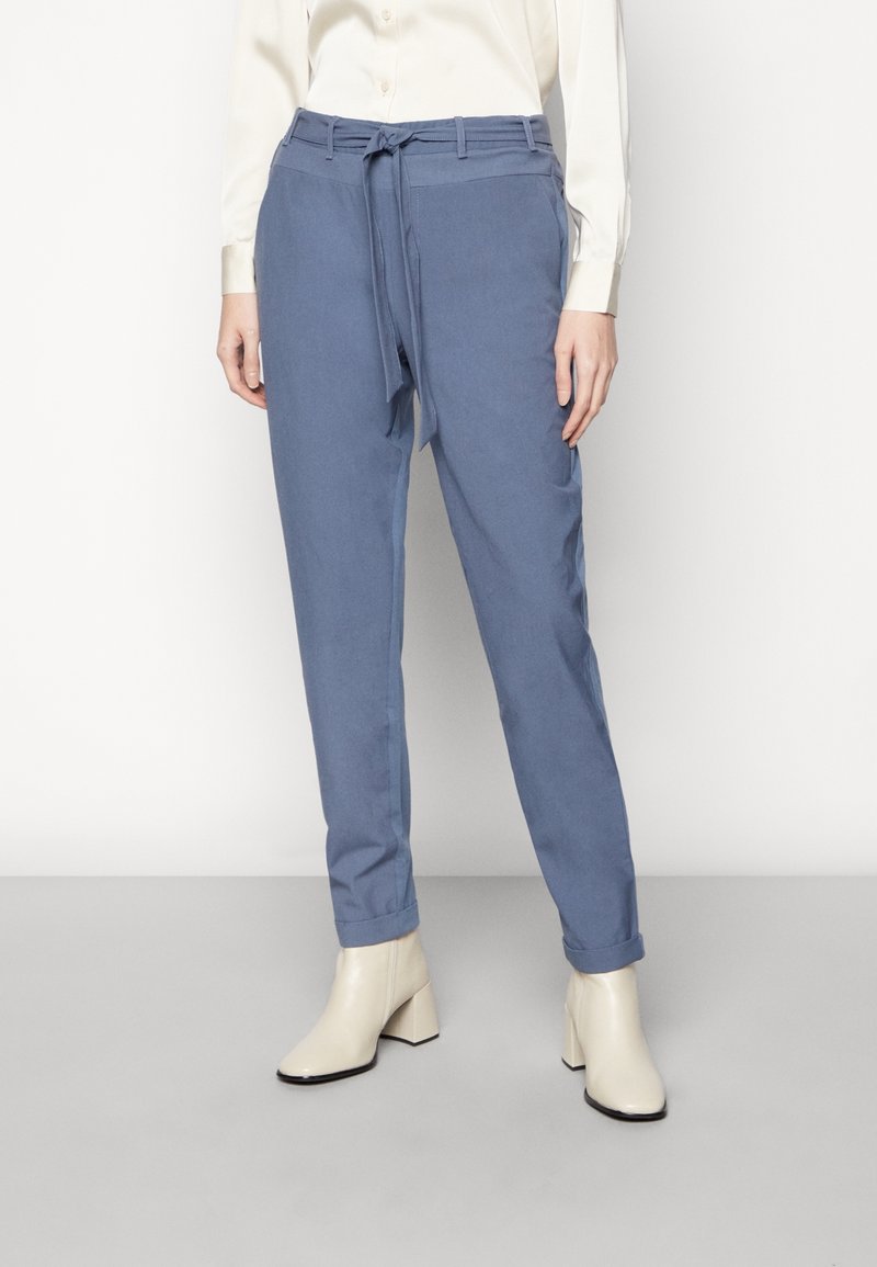 Femme portant un pantalon bleu clair taille haute avec un nœud à l'avant, associé à un chemisier crème et des bottines blanches à talons, debout sur un sol gris.