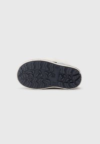 Suela de goma negra de un zapato infantil con surcos profundos y un diseño en la suela, y bordes de entresuela blanca sobre un fondo blanco liso.