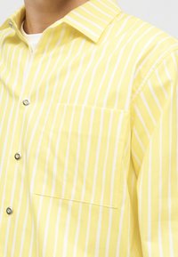 Gelbes Hemd mit weißen vertikalen Streifen, einer Brusttasche und Kragen, getragen über einem weißen Unterhemd.