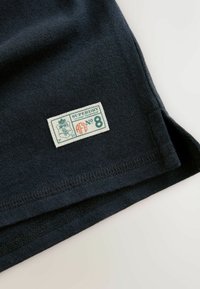 Țesătură bleumarin închis cu o etichetă albă dreptunghiulară cusută pe care scrie "SUPERDRY No 8" și un mic emblemă în colțul de jos.