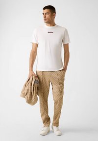 T-shirt en coton blanc avec logo "GUESS" rouge, pantalon beige à motifs, tenant un pull beige, et portant des baskets blanches.