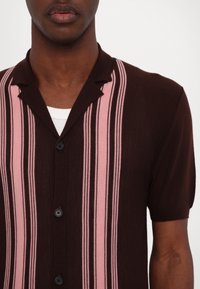 Košile s krátkým rukávem z tmavě hnědé látky, která má vertikální růžové pruhy, límec na knoflíky a černé knoflíky na přední straně.