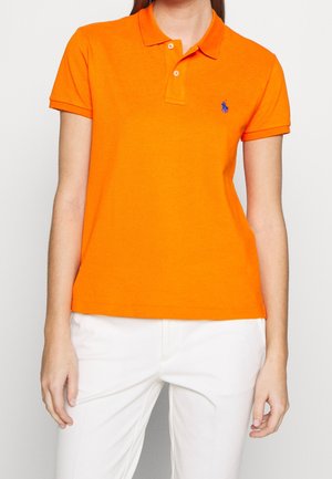 Vrouw draagt een feloranje polo met korte mouwen en een blauw logo, gecombineerd met een witte broek, tegen een effen achtergrond.