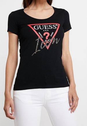 Vrouw draagt een zwart Guess T-shirt met een rood driehoeklogo en de tekst "Icon", gecombineerd met een witte broek, staand tegen een effen achtergrond.