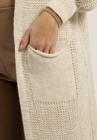 Long cardigan beige avec un design en tricot texturé. Il dispose d'une grande poche avant et de poignets côtelés, présenté sur un bras tenant la poche.