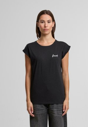 Femme aux cheveux bruns et raides porte un t-shirt noir à manches retroussées avec le texte "forever" sur la poitrine et un jean gris foncé, debout devant un fond gris.