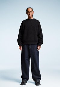 Czarny sweter z dzianiny z prążkowanym dekoltem, zestawiony z luźnymi ciemnymi dżinsami i ciemnymi trampkami. Model stoi na jasnym tle.