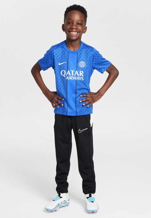 Blå PSG-drakt med geometrisk mønster, svarte Nike-bukser og hvite fotballsko med blå detaljer. Barn smiler og poserer selvsikkert.