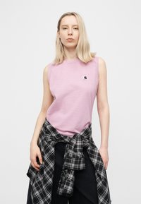 Νεαρή γυναίκα με ξανθά μαλλιά, που φοράει ένα ροζ αμάνικο τοπ και ένα μαύρο-λευκό καρό πουκάμισο δεμένο γύρω από τη μέση της.