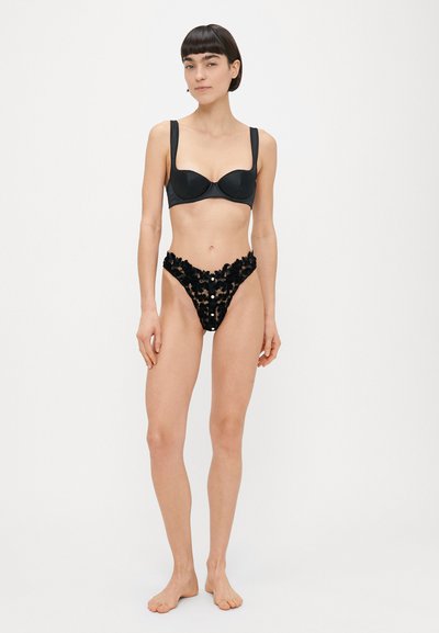 Femme debout pieds nus portant un soutien-gorge noir à armatures et une culotte taille haute en dentelle florale noire sur un fond blanc uni.