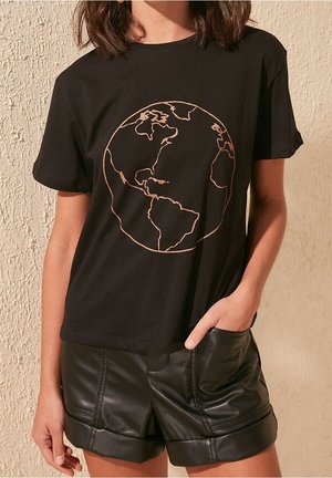 Woman wearing black T-shirt with simple earth outline graphic and black leather shorts, standing against textured beige wall.