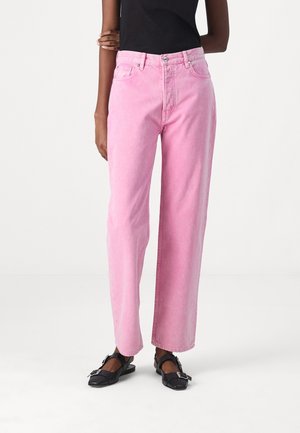 Person iført højtaljede, straight-leg pink jeans og sorte spidse remsko med spænde, stående mod en ensartet hvid baggrund.