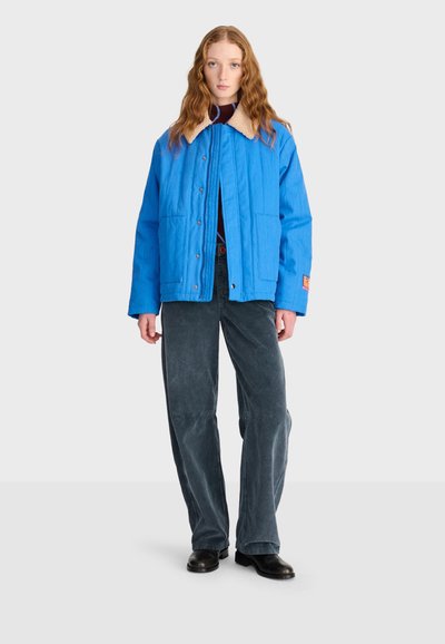 Jeune femme aux longs cheveux ondulés portant une veste bleu vif avec un col beige, un pantalon large gris foncé et des chaussures noires, debout, droite.