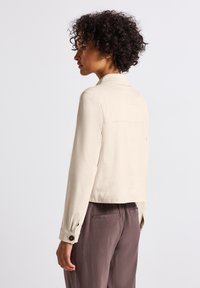 Beige kort jacka med krage, knäppta manschetter och en slät yta. Bärs över gråa byxor, vilket visar en enkel, ren design.