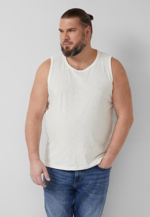 Brodaty mężczyzna z włosami związanymi do tyłu, ubrany w białą koszulkę bez rękawów i niebieskie jeansy, stoi z jedną ręką w kieszeni.