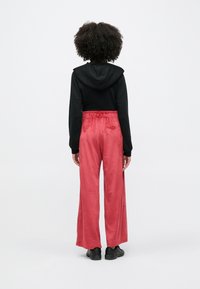 Osoba s kudrnatými vlasy, oblečená v černé mikině s kapucí, širokých růžových sametových kalhotách a černých teniskách, stojící zády k pozorovateli na bílém pozadí.